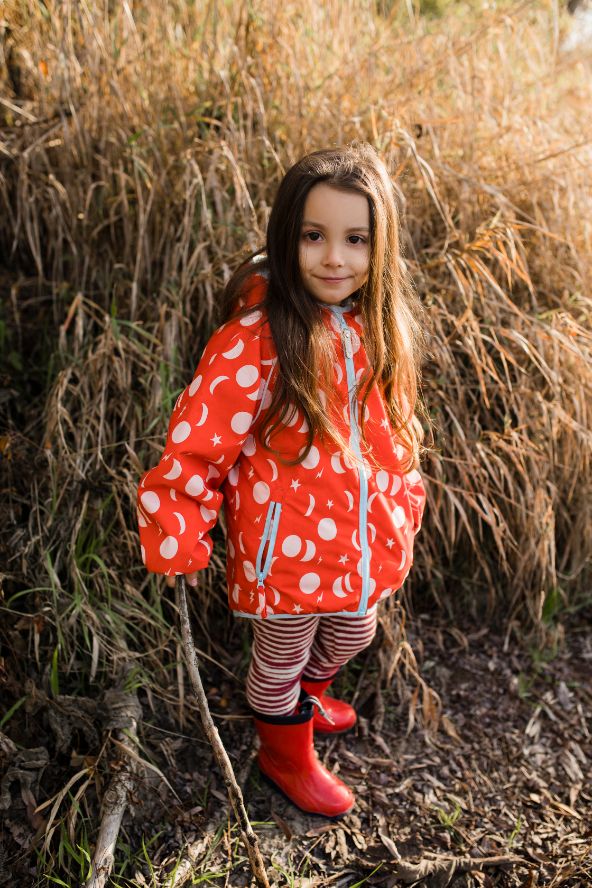 Bambina in piedi indossa giacca impermeabile rossa con stelle e luna, leggings a righe panna e rossi, rivaletti rossi