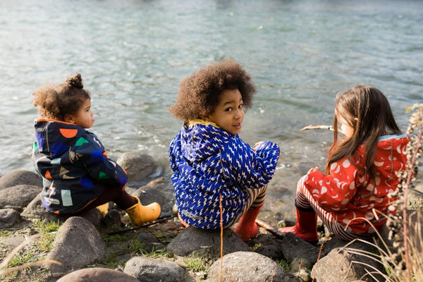Tre bambini seduti in riva al fiume indossano giacche impermeabili colorate