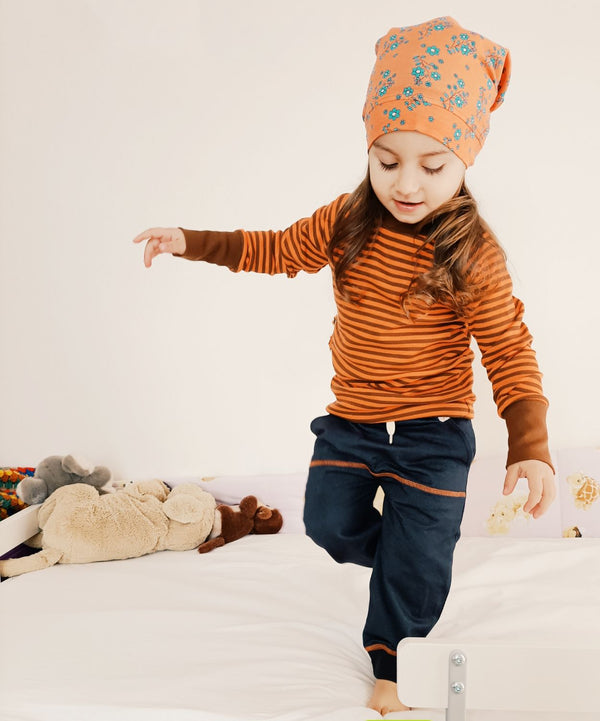 Bambina indossa cuffietta arancione a fiori, maglietta a maniche lunghe arancione con righe marroni e pantaloni lunghi grigi con dettagli arancioni