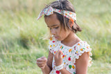 bambina in primo piano gioca in un campo con un filo d'erba indossa un abito color panna con alberi di mele disegnati e fascia in testa a fiori