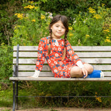 bambina seduta su una panchina indossa abito rosso con fiori bianchi e blu. alle sue spalle fiori gialli