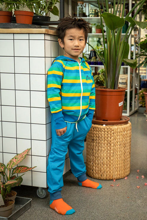 bambino appoggiato ad una parete indossa felpa azzurra a righe gialle e blu e pantaloni blu