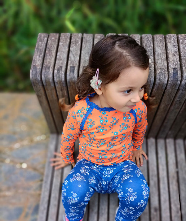 bambina seduta indossa maglietta a maniche lunghe arancione con fiorellini e leggings blu con fiorellini