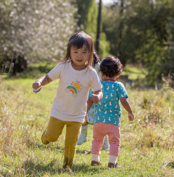 bambina corre in un prato con maglietta bianca con arcobaleno maniche corte e pantaloni gialli
