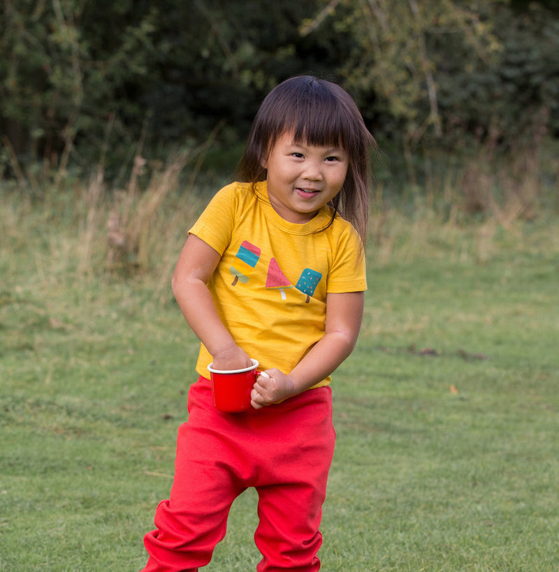 bambina con in mano una tazza rossa indossa maglietta gialla conm stampa colortata maniche corte in cotone pantaloni rossi